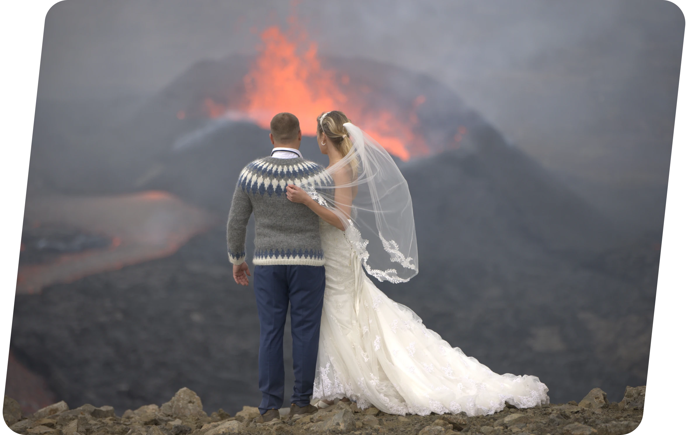 A bride in a white wedding dress and veil stands beside a groom wearing a patterned sweater and blue trousers, both facing an erupting volcano with glowing lava in the background.