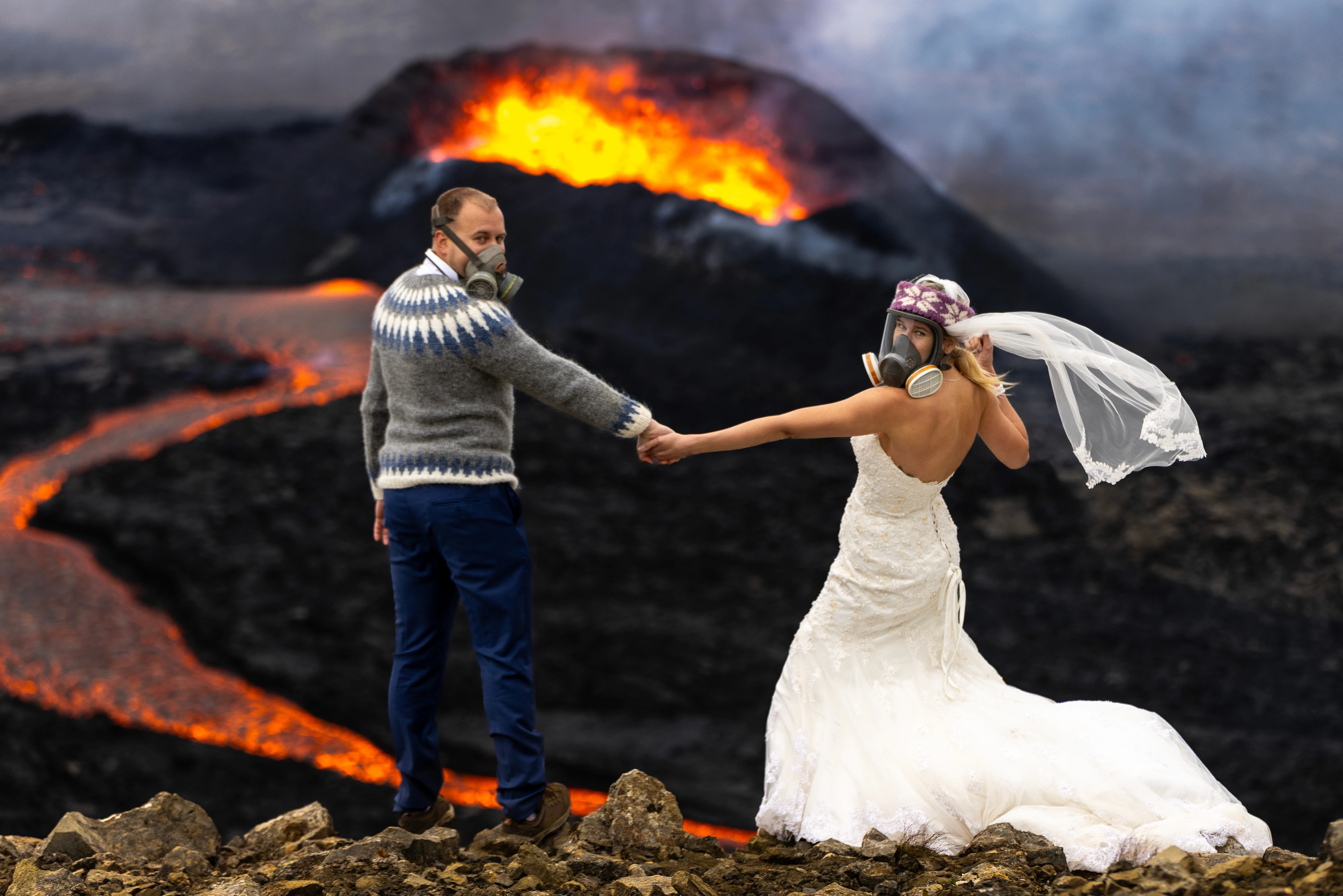 A bride and groom in formal wedding attire wear gas masks while holding hands in front of an erupting volcano, with flowing lava behind them.