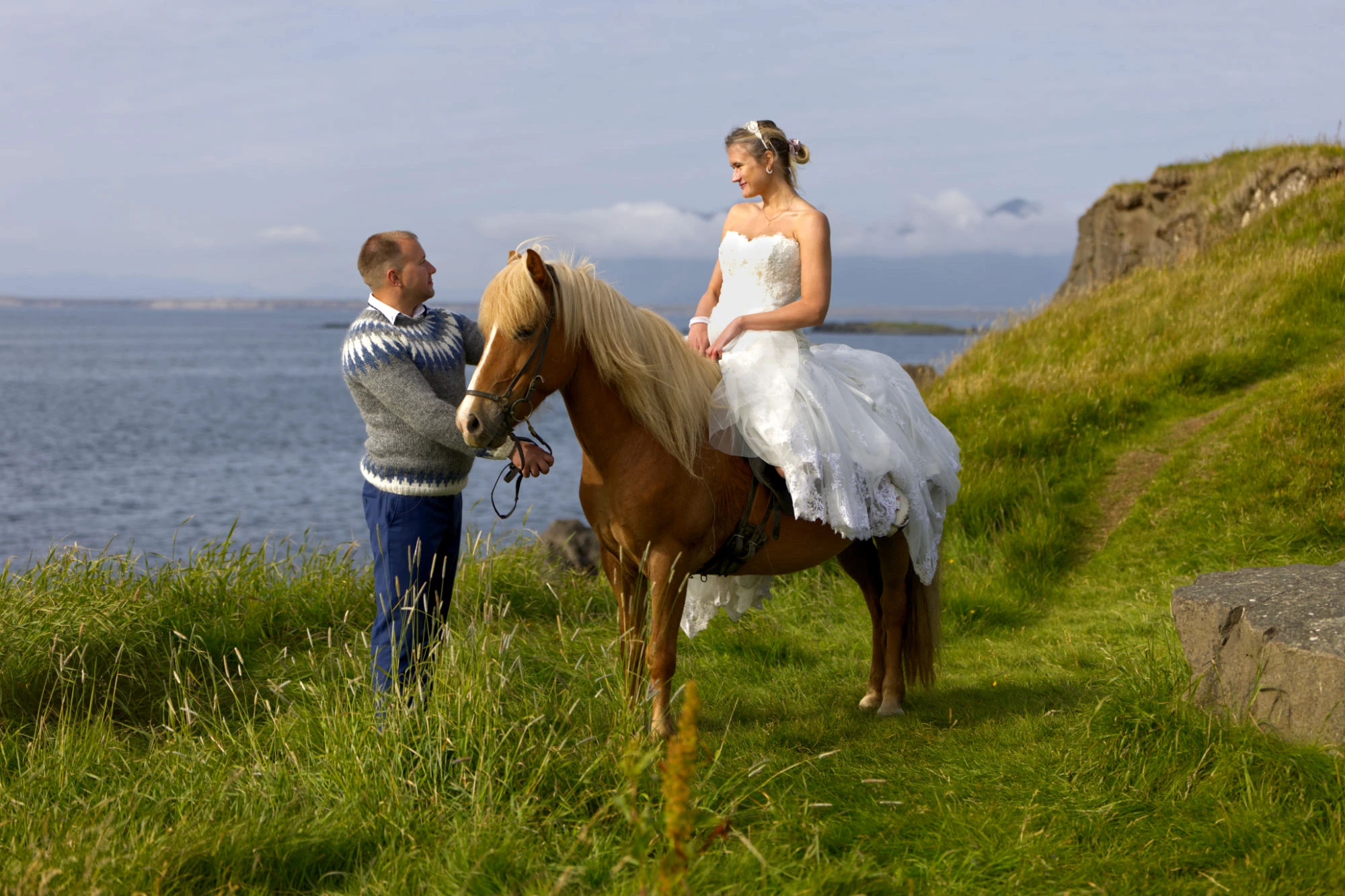 A bride in a white dress rides an Icelandic horse, led by a groom through grassy hills under a bright sky.